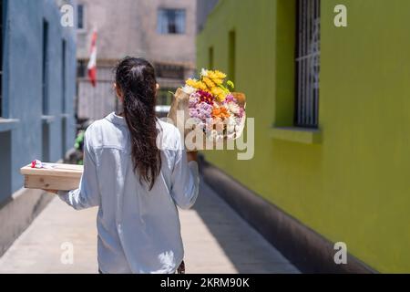 Back view of unrecognizable female in casual outfit walking on street with bouquet of flowers wrapped in craft paper and gift box Stock Photo