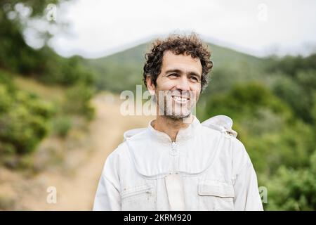Positive bearded male with curly hair wearing beekeeper suit while standing in forest smiling and looking away Stock Photo