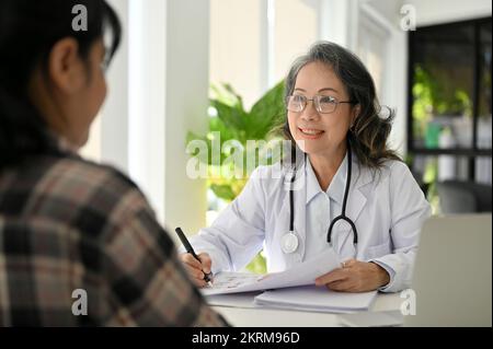 Happy and cheerful Asian aged female doctor having a nice conversation with her patient in the clinic during medical checkup. Stock Photo