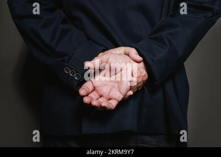 Crop unrecognizable aged male in black jacket seen from behind with hands clasped behind back on gray background in studio Stock Photo