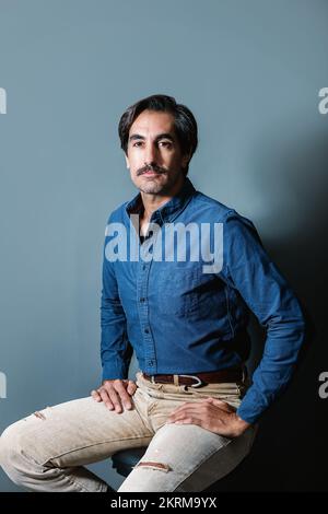 Confident young male in blue shirt and jeans sitting on gray background and looking at camera Stock Photo