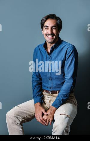 Happy young male in blue shirt and jeans sitting on gray background and looking at camera Stock Photo
