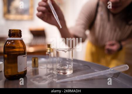 Cropped unrecognizable craftswoman dripping essential oil with pipette into flask while standing near table in blurred room during perfume preparation Stock Photo