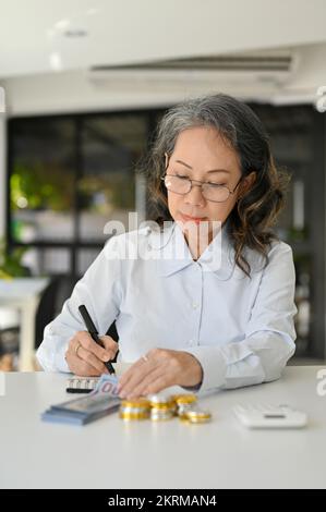 Portrait, Professional Asian aged female accountant using calculator, calculating and managing financial sales accounts, working at her desk. Stock Photo