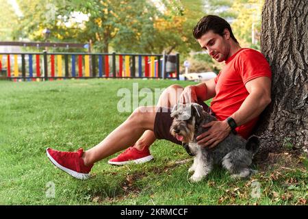 Side view full body of adult male athlete in red sportswear sitting near tree with cute Miniature Schnauzer during walk in park Stock Photo