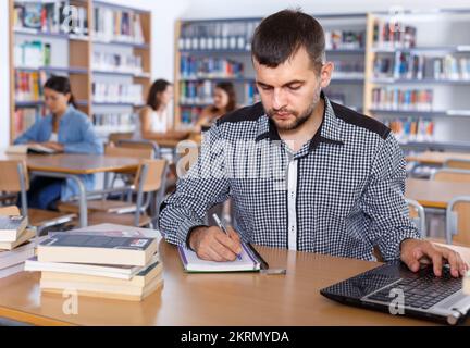 Young focused man student working with laptop and books in public library Stock Photo