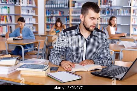 Focused guy preparing for exam in college library, reading book and using laptop for searching information Stock Photo