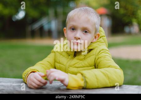 A boy sits at a table in the park in the fall season. Child Boy Son In Autumn Park, Sitting On Wooden Bench And Table. Little Kid Outdoors. Stock Photo