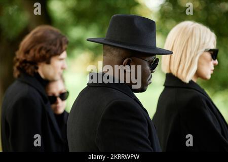 Side view of people wearing all black with sunglasses standing in silence at outdoor funeral ceremony Stock Photo