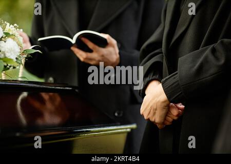 Close up of young woman standing by coffin at outdoor funeral ceremony, priest reading prayer in background, copy space Stock Photo
