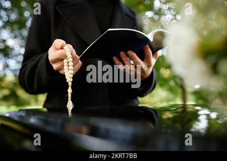 Close up of priest holding bible and rosary reading prayer by coffin at outdoor funeral ceremony, copy space Stock Photo