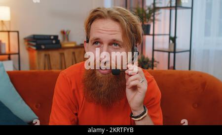 POV of bearded man wearing headset, freelance worker, call center or support service operator helpline, having talk with client or colleague communication support waving hello alone at home couch Stock Photo