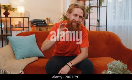 Ok. Happy cheerful bearded ginger man looking approvingly at camera showing ok gesture, positive like sign, approve something good. Young caucasian redhead guy at home in room sitting on orange couch Stock Photo