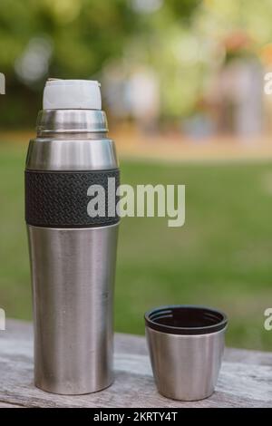 Stainless steel cup, mug with thermos stands on a wooden table in the forest on a green natural background. Thermos and mug with hot drink standing on Stock Photo