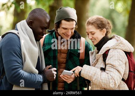 Diverse group of friends using smartphone and smiling while hiking outdoors Stock Photo