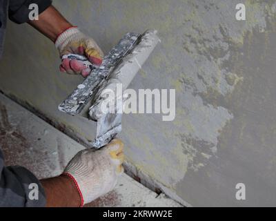 hands of a plasterer in fabric gloves with two spatulas while working with cement mortar in the process of plastering walls in a room Stock Photo