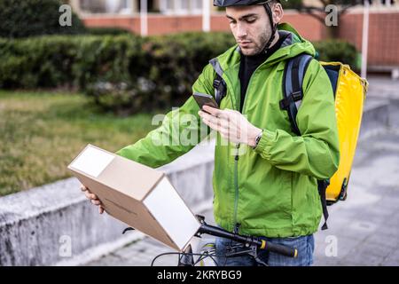 handsome young man using bicylce to deliver boxes, documents and food at home, male courier using technology of smart devices to find address and send Stock Photo