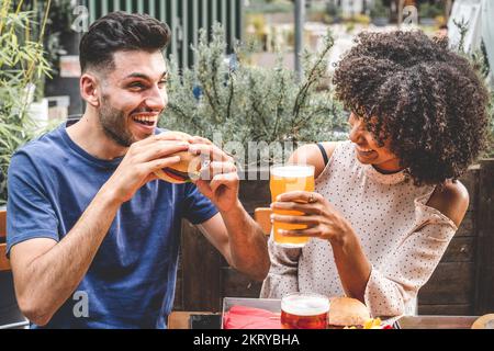 young couple having fun at restaurant, two diverse friends eating burgers at fast food and drinking beer Stock Photo