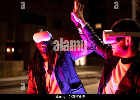 Young multiracial friends playing in virtual reality and win, people giving five to eac other with ar googles, neon light effect, nft transaction succ Stock Photo