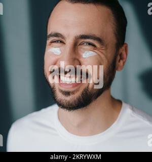 Portrait of a cheerful male looking at camera with smile and having cream on his cheeks, during skin care routine against studio background. Natural Stock Photo