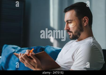 Close-up portrait of a young man with eye-patches sitting on sofa writing in diary. Modern lifestyle. People lifestyle. Stock Photo