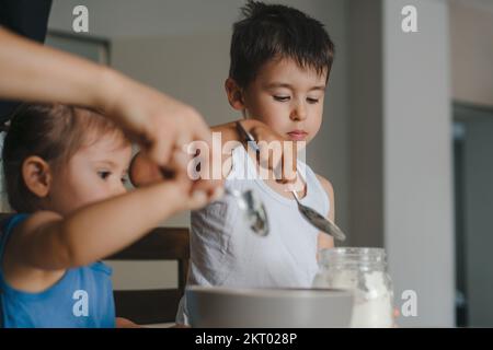 Family with adorable two little kids cooking in modern kitchen pancakes together. Cake mix preparation, make yummy home-made dessert, enjoy Stock Photo
