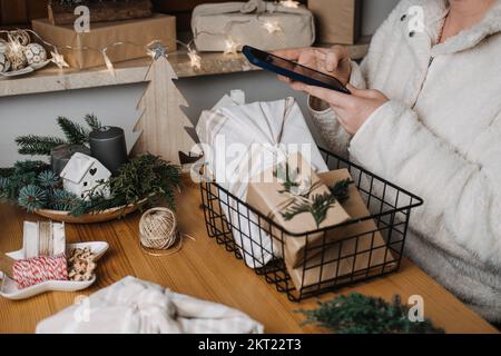 Woman with smartphone in hand and Christmas eco friendly wrapping gifts on table. Woman using cell phone near gifts in craft paper on table at home Stock Photo
