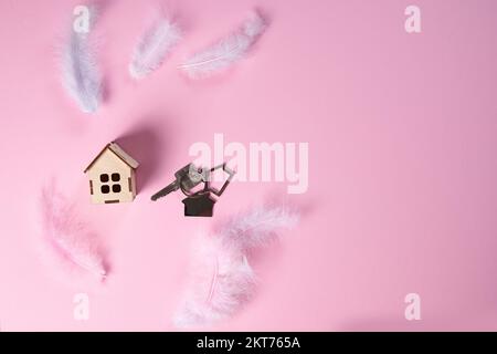 A wooden house and the keys on a pink pastel background with feathers. The concept of an easy loan, mortgage, insurance, copy space for your advertising. Flat lay. Stock Photo