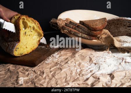 Men's hands cut a loaf of bread on a cutting board on a dark black background. Copy space.The concept of ecology, home baking, small business. Coffee or bakeries. Stock Photo