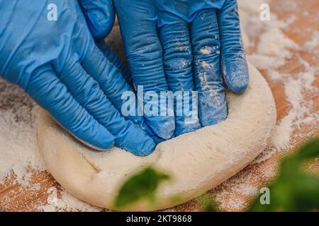 Hands with plastic gloves preparing dough for pizza on a wooden board with white flour and ingredients around, close up Stock Photo