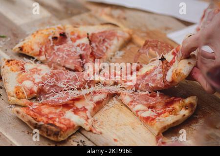 Hands taking pieces or slices of pizza with tomato sauce, mozzarella, parmesan cheese and salami meat from a wooden board Stock Photo