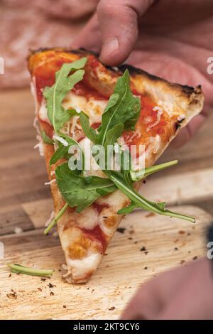 Hand taking last piece or slice of pizza with tomato sauce, mozzarella and fresh rocket leaves from a wooden board, close up, vertical Stock Photo