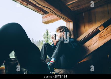 Young teenage boy with hot tea cup drinking while he sitting on the wooden forest house terrace and enjoying the morning spruce foggy forest. Travelin Stock Photo