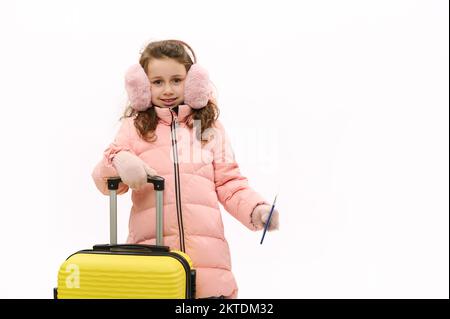 Cute little girl wearing pink fluffy earmuffs, down jacket and wool mittens, with suitcase and boarding pass, on white Stock Photo