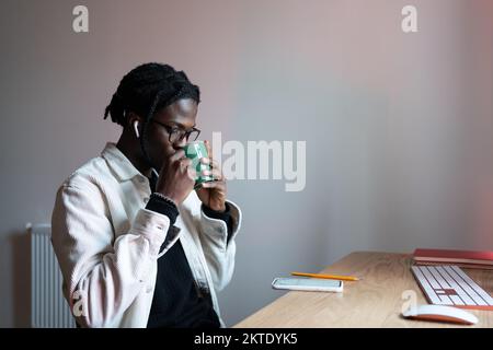 African student guy sitting at desk drinking tea waiting for webinar to start while studying at home Stock Photo