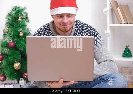 Man with laptop working, studying remotely new year 2023, Christmas. Man in Santa hat on background Christmas tree, garlands, gifts. Remote work. Surfing Internet. Man congratulates on holiday online. Stock Photo