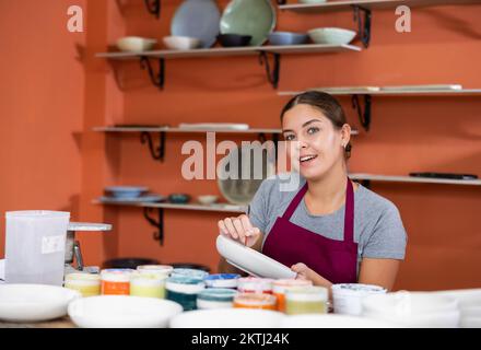 Woman potter with paintbrush glazing, painting on plate in workshop, working in pottery Stock Photo