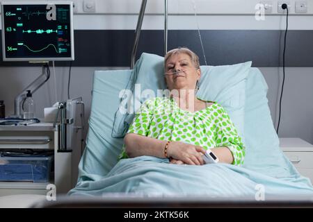 Female patient resting after surgery in hospital monitoring room. Elderly lady connected to medical equipment measuring vital signs. Old woman relaxing in sanatorium recovery bedroom. Stock Photo