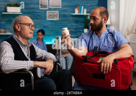 Elderly man seated in wheelchair receiving bottle of pills from nurse for health treatment and recovery. Specialist giving a bottle of medication to patient with disability in nursing home. Stock Photo