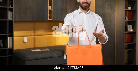 Unrecognizable male retailer in shirt giving orange shopping bag to camera in furniture store. Crop view of smiling bearded seller, offering purchase, with cupboard on background. Concept of shopping. Stock Photo
