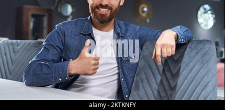 Unrecognyzable bearded man showing thumb up, while sitting at table in cafe. Crop view of smiling male in casual clothes gesturing like sign, while resting indoors. Concept of approval hand gesture. Stock Photo