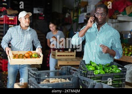 Owner of vegetable warehouse is talking on mobile phone while other workers are sorting vegetables Stock Photo