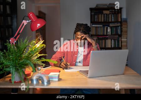 Hardworking young African guy student writing notes while watching webinar on laptop at home Stock Photo