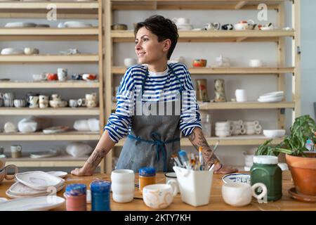 Calm female ceramist with tattoos in pottery workshop looking at window and smiling, plans projects. Stock Photo