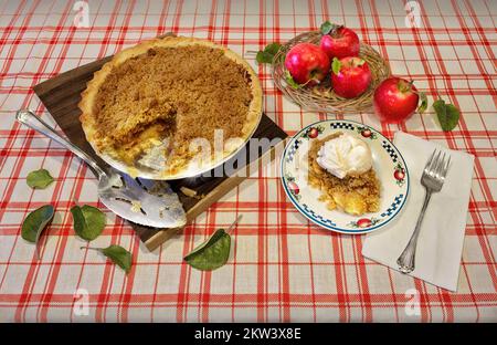 Homemade organic apple pie dessert ready to eat. Slice of pie with ice cream on top, freshly picked apples in a basket, on red and white tablecloth. Stock Photo