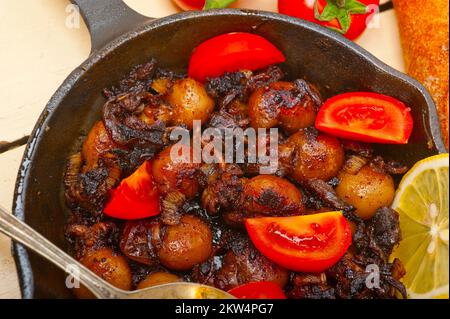 Baby cuttle fish roasted on iron skillet with tomatoes and onions over rustic wood table Stock Photo