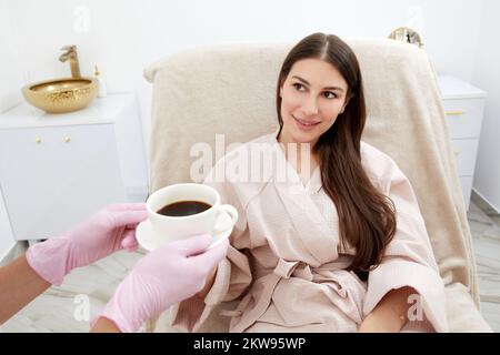 Beautician serving cup of coffee to her client after body treatment in beauty salon. Relaxing with beverages on a cosmetic bed. Beauty , pleasure Stock Photo