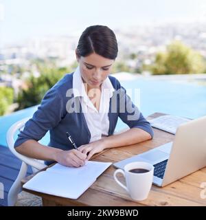Taking notes. a young businesswoman working on her laptop by a swimming pool. Stock Photo