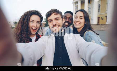 Point of view shot of multiracial group of tourists taking selfie in city center holding gadget camera and posing together with hand gestures expressing positive emotions. Stock Photo