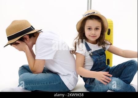 Little girl wearing straw hat, sits near her brother who hides his face, against yellow suitcase and looking at camera Stock Photo